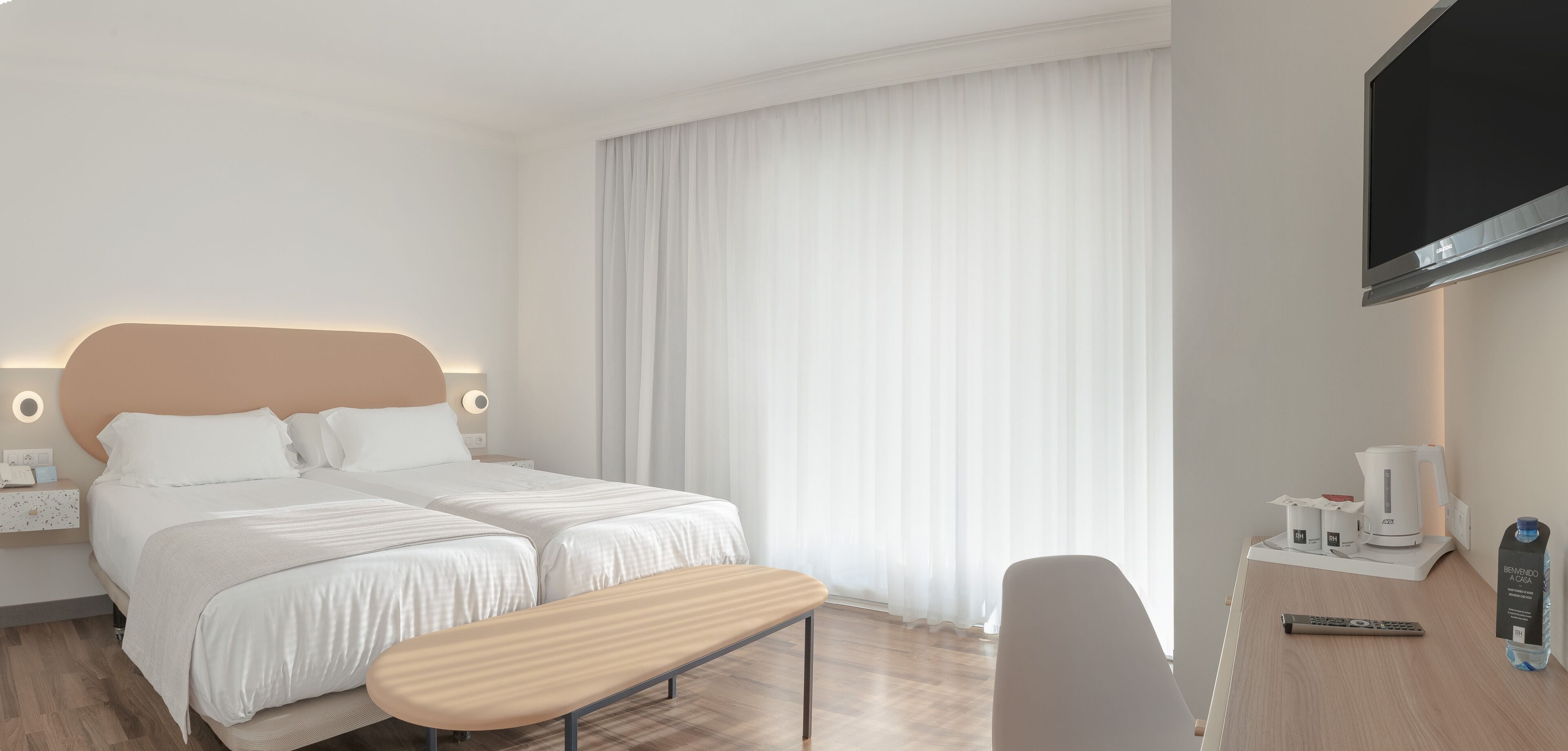 Habitación de hotel con cama doble, mesita de noche con lámpara, televisor, cafetera y botella de agua.