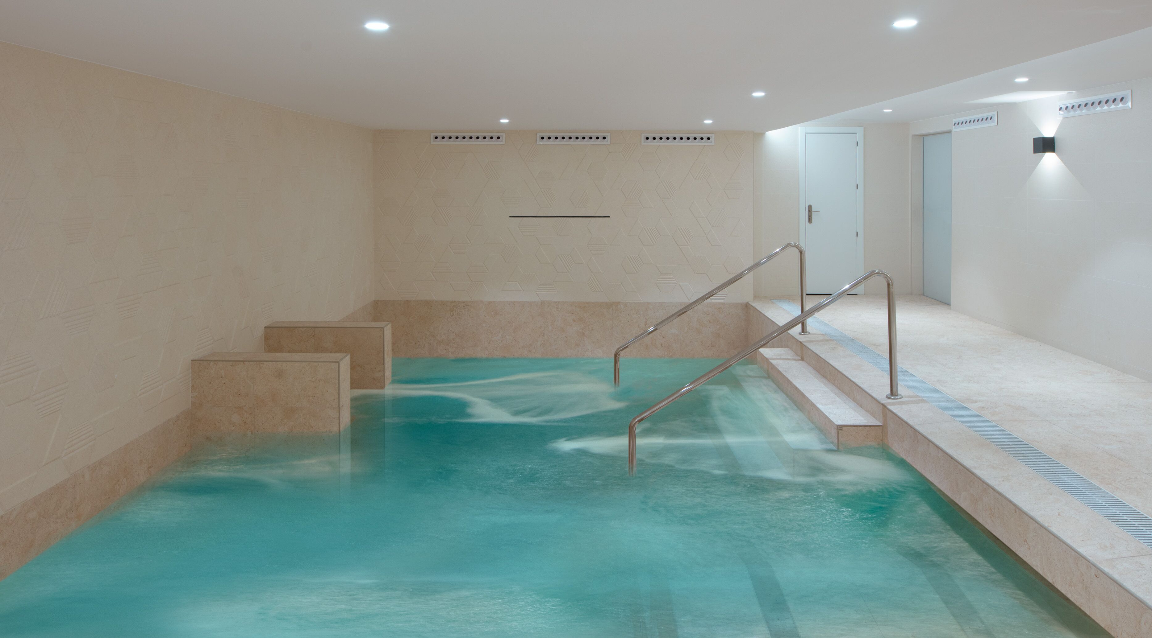 An indoor swimming pool with clear water, steps leading into the pool, and a tiled wall with a geometric pattern.
