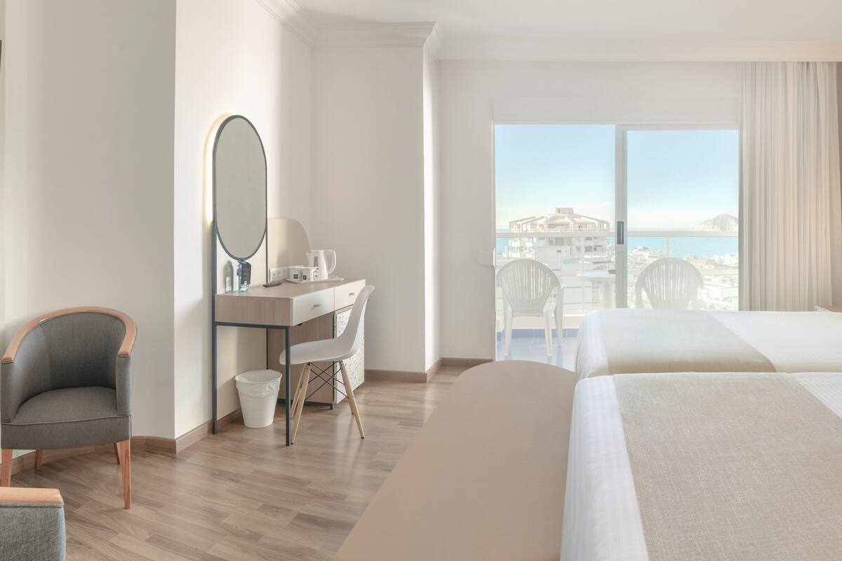 A hotel room with two beds, a desk with a chair, a mirror, and a view of the sea.