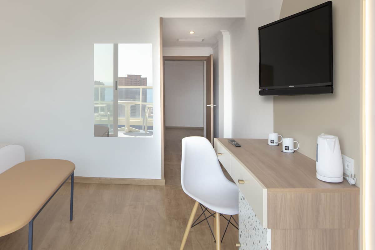 A hotel room with a bed, bedside tables, a desk with a chair, a flat-screen TV, and a window with a view of buildings.