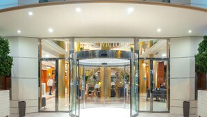 Entrada de hotel con puerta giratoria, paredes de vidrio y una escalera visible que conduce a un nivel superior.