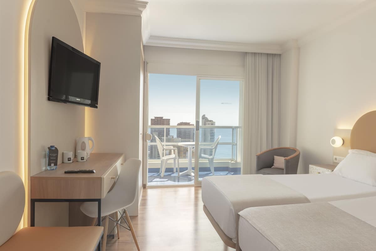 A hotel room with two beds, a desk with a TV, a chair, a balcony with a view of buildings, and a phone on the nightstand.
