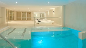 Un área interior de piscina moderna con barandillas de vidrio transparente, una zona de estar con tumbonas y una entrada visible a otra habitación.
