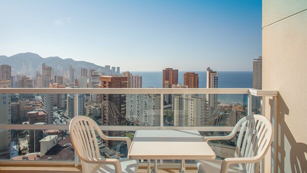 Un balcón con una mesa y dos sillas, con vistas a un paisaje urbano que incluye edificios y una masa de agua.