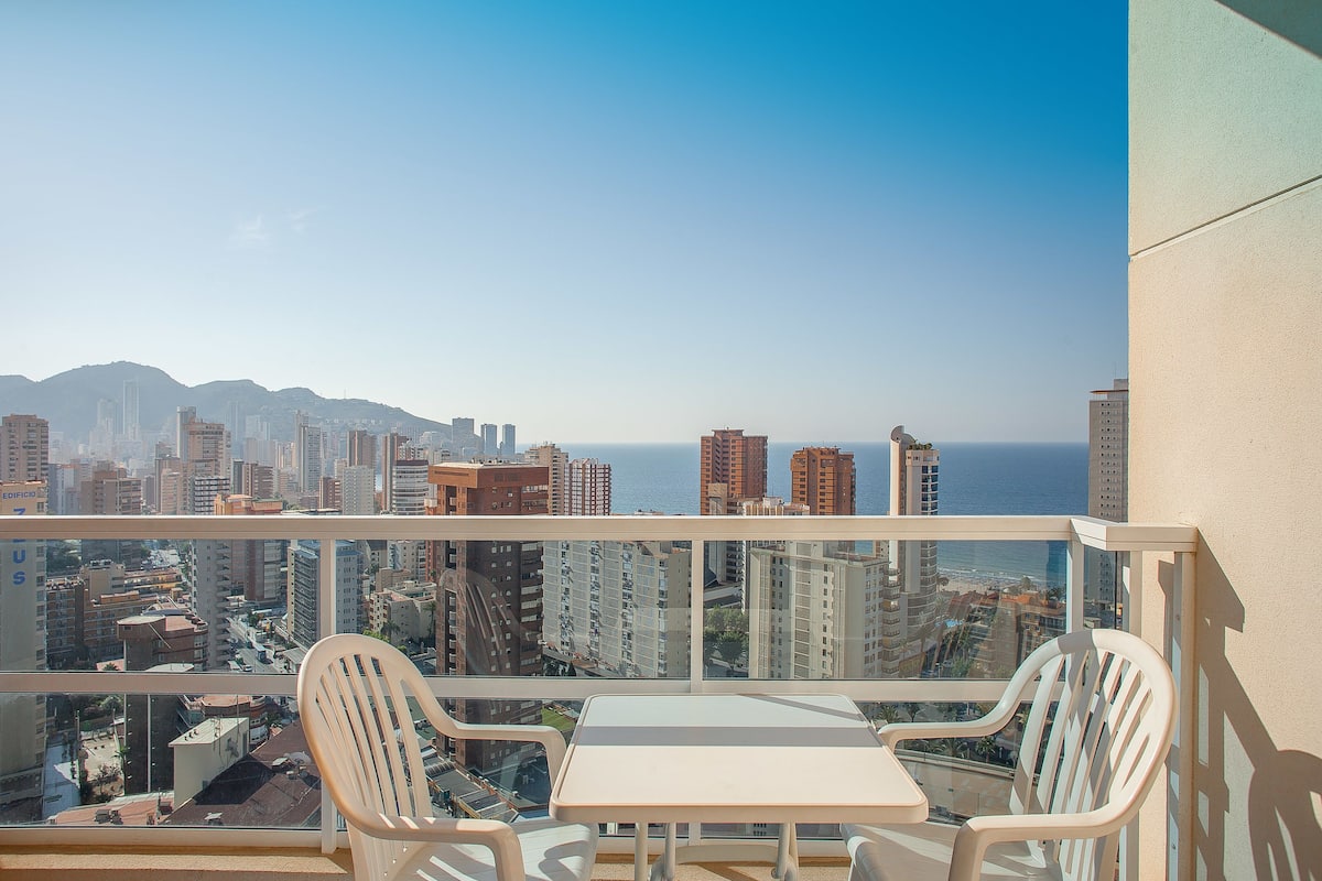 A balcony with a table and chairs, overlooking a cityscape and the ocean.