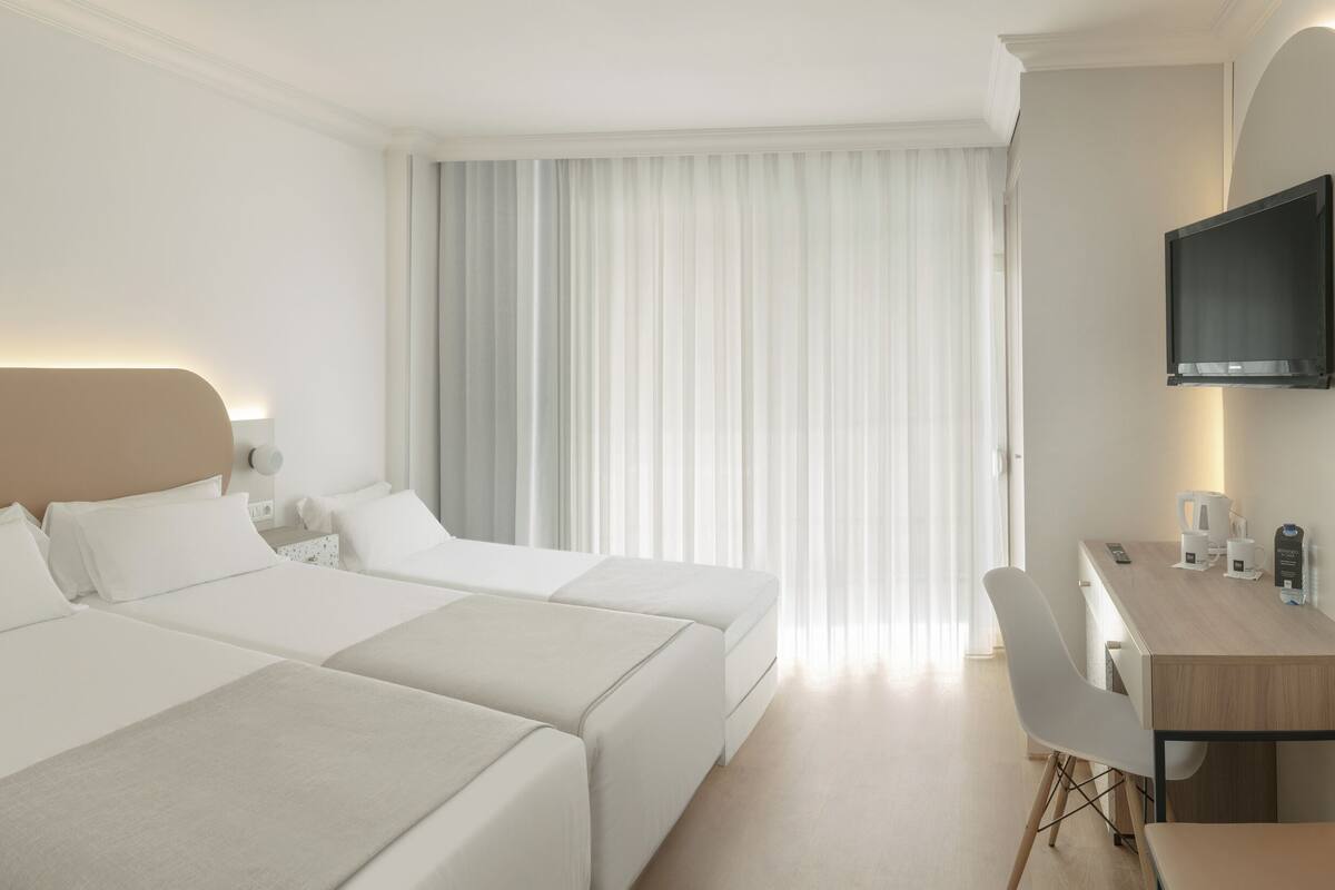 A hotel room with two beds, a desk with a chair, a TV mounted on the wall, and a window with curtains.