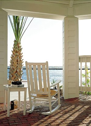 A white rocking chair and a table with a plant and a book, positioned near a white door with a view of the ocean. - Lighthouse Inn At Aransas Bay (Rockport)