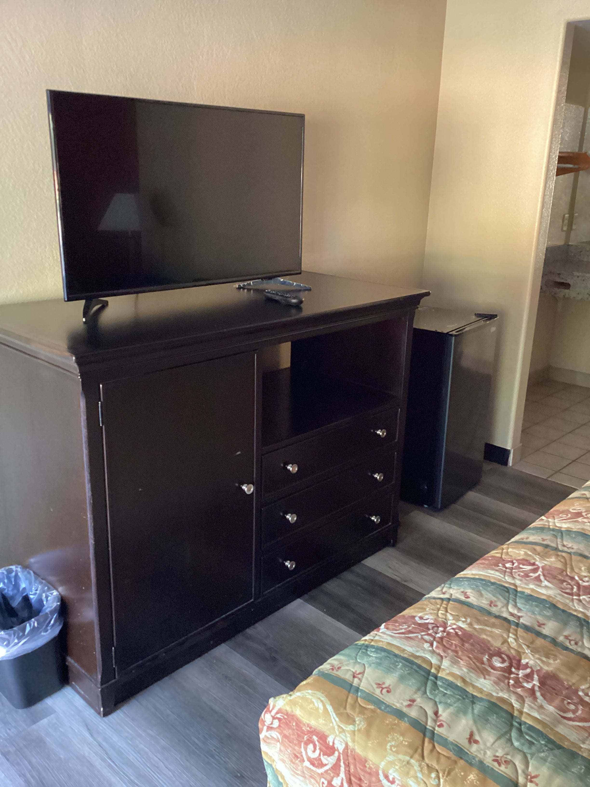 Une pièce avec un meuble en bois foncé, un téléviseur posé dessus, une poubelle et un lit recouvert d’une couvre-lit à motifs.