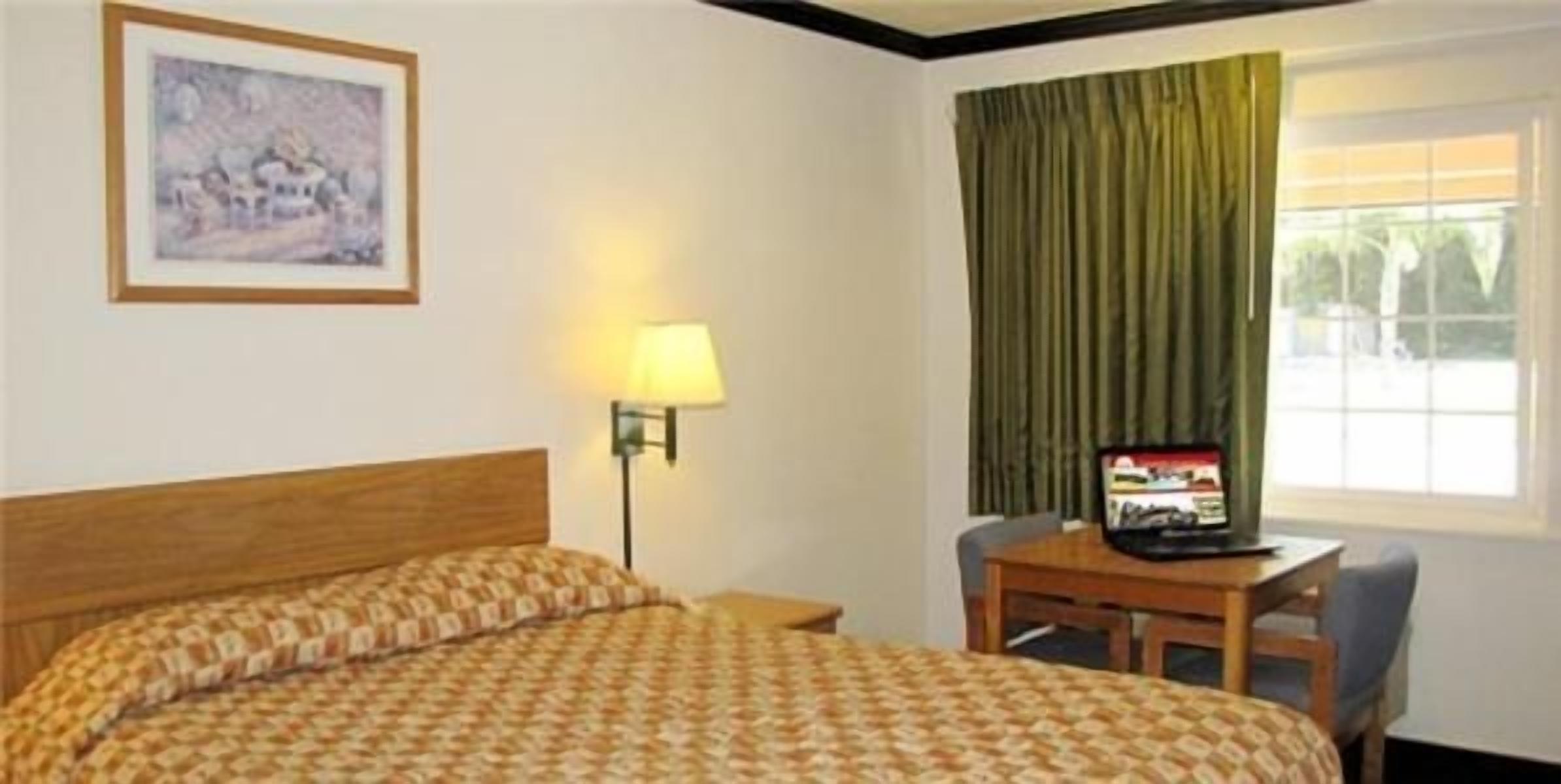 Habitación de hotel con cama, escritorio con silla, lámpara, televisor y ventana con cortinas.
