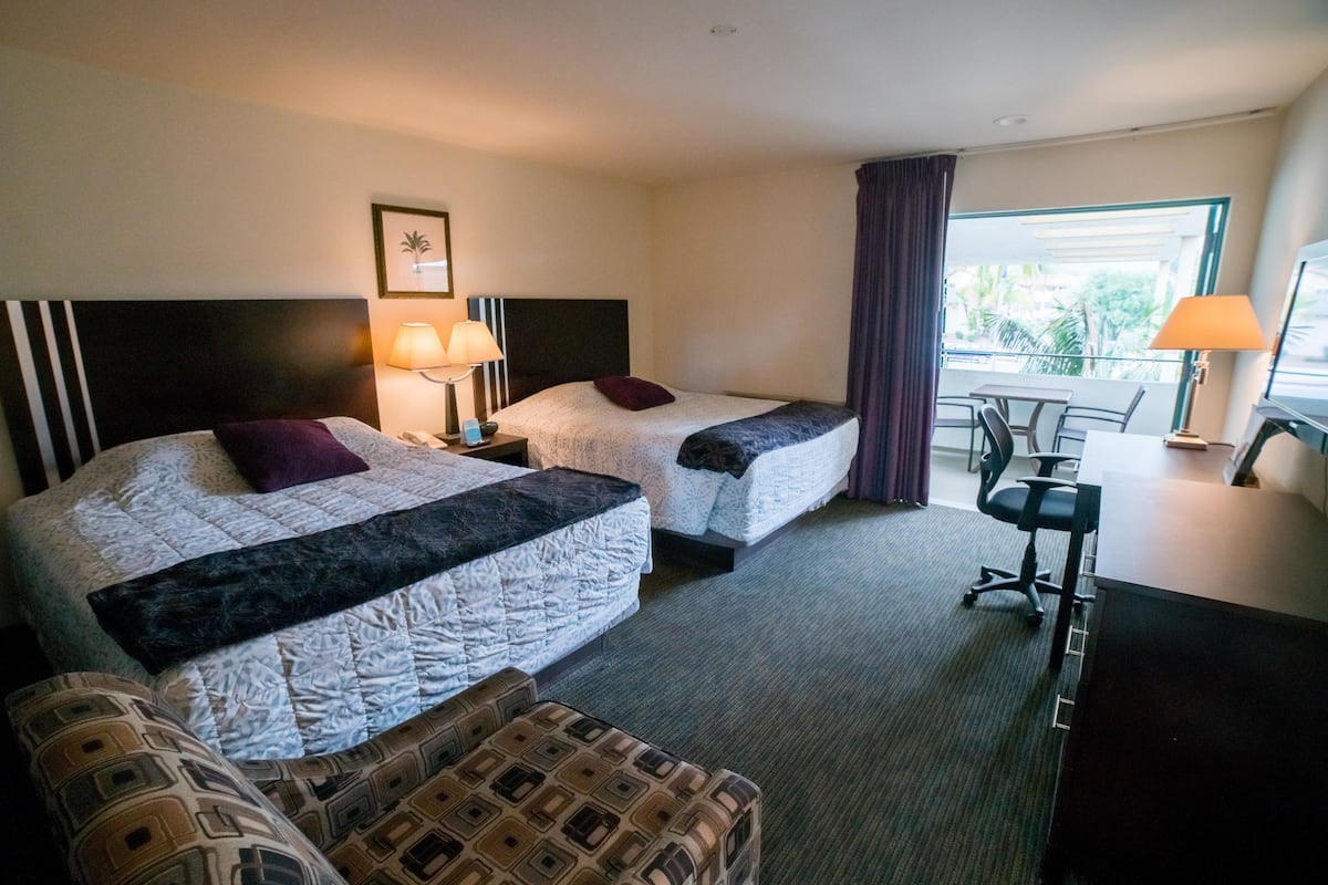 A hotel room with two beds, a desk, a chair, and a window with a view of palm trees.