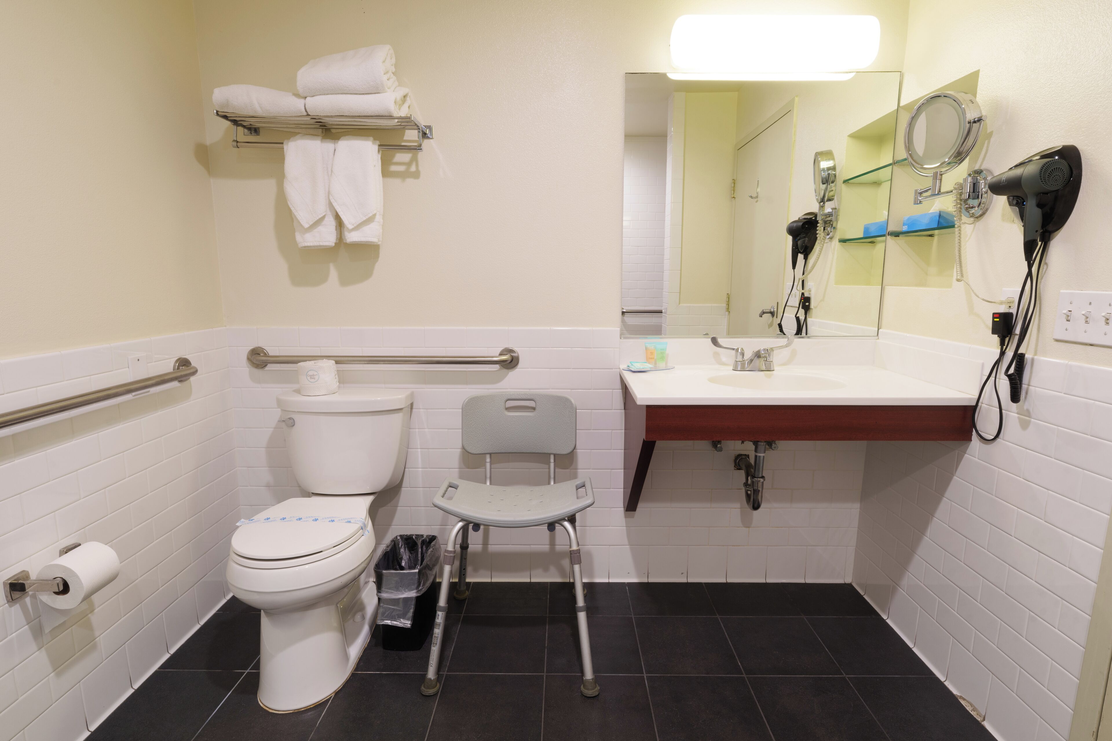 Une salle de bain équipée de toilettes, d’un sèche-mains, d’un lavabo, d’un miroir et d’une barre d’appui.