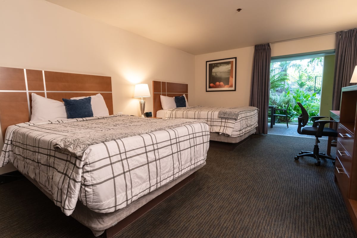 A hotel room with two beds, a desk, a chair, a lamp, and a window with a view of greenery.
