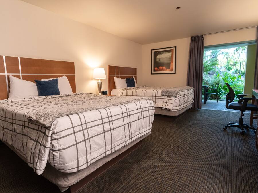 Ett hotellrum med två sängar, ett skrivbord, en stol, en lampa och ett fönster med utsikt över grönområden.