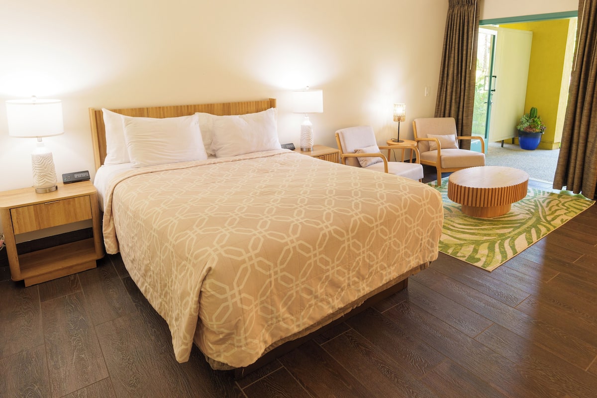 A hotel room with a large bed, two bedside tables, a chair, a small table, and a potted plant.