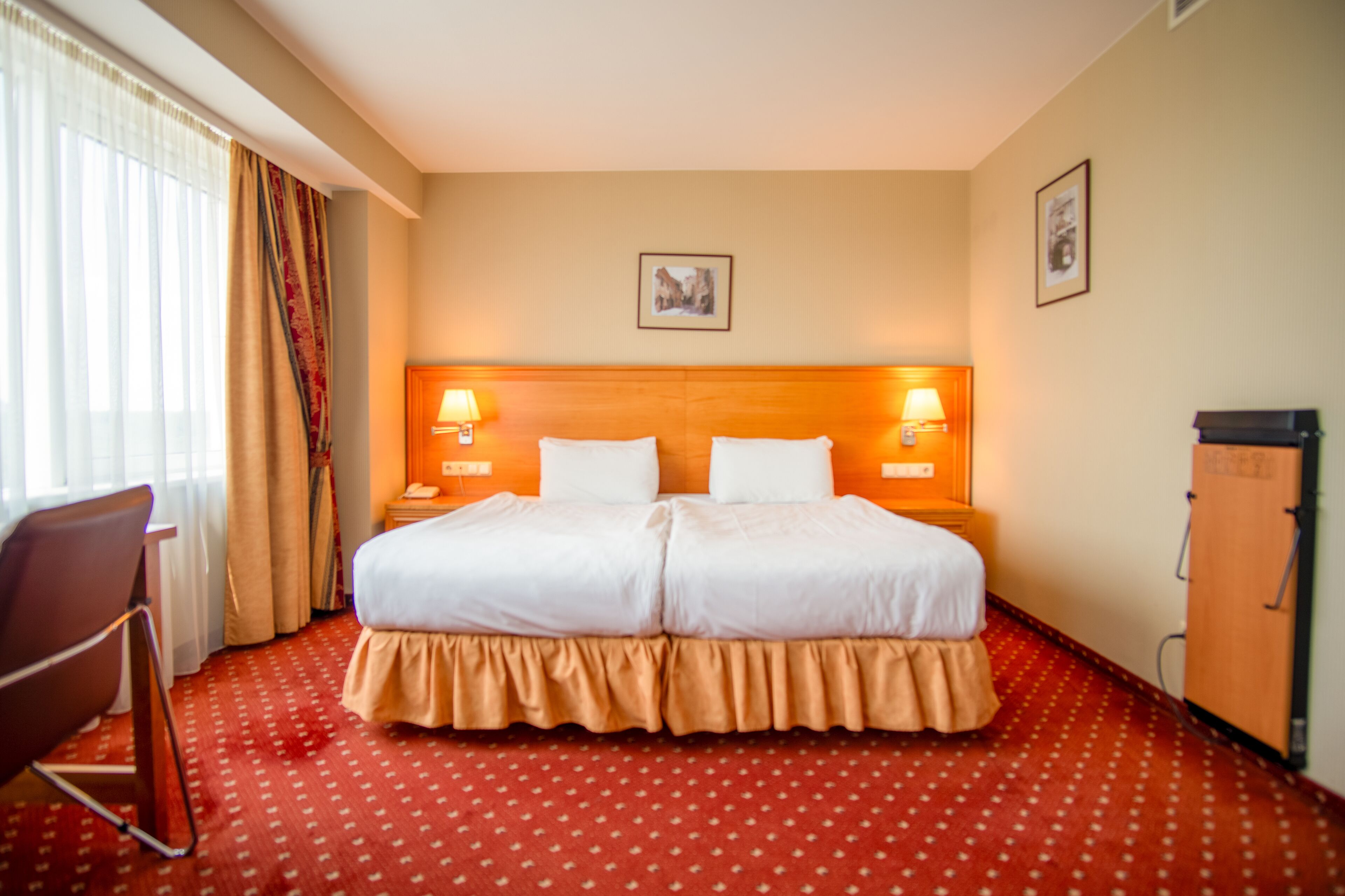 A hotel room with a double bed, two bedside lamps, a wooden headboard, a red patterned carpet, a wooden wardrobe, and a chair.