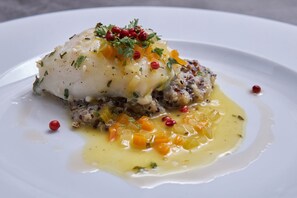 Ein weißer Teller mit einem Fischfilet, belegt mit Kräutern und roten Pfefferkörnern, dazu eine gelbe Soße und gewürfelte Gemüse.