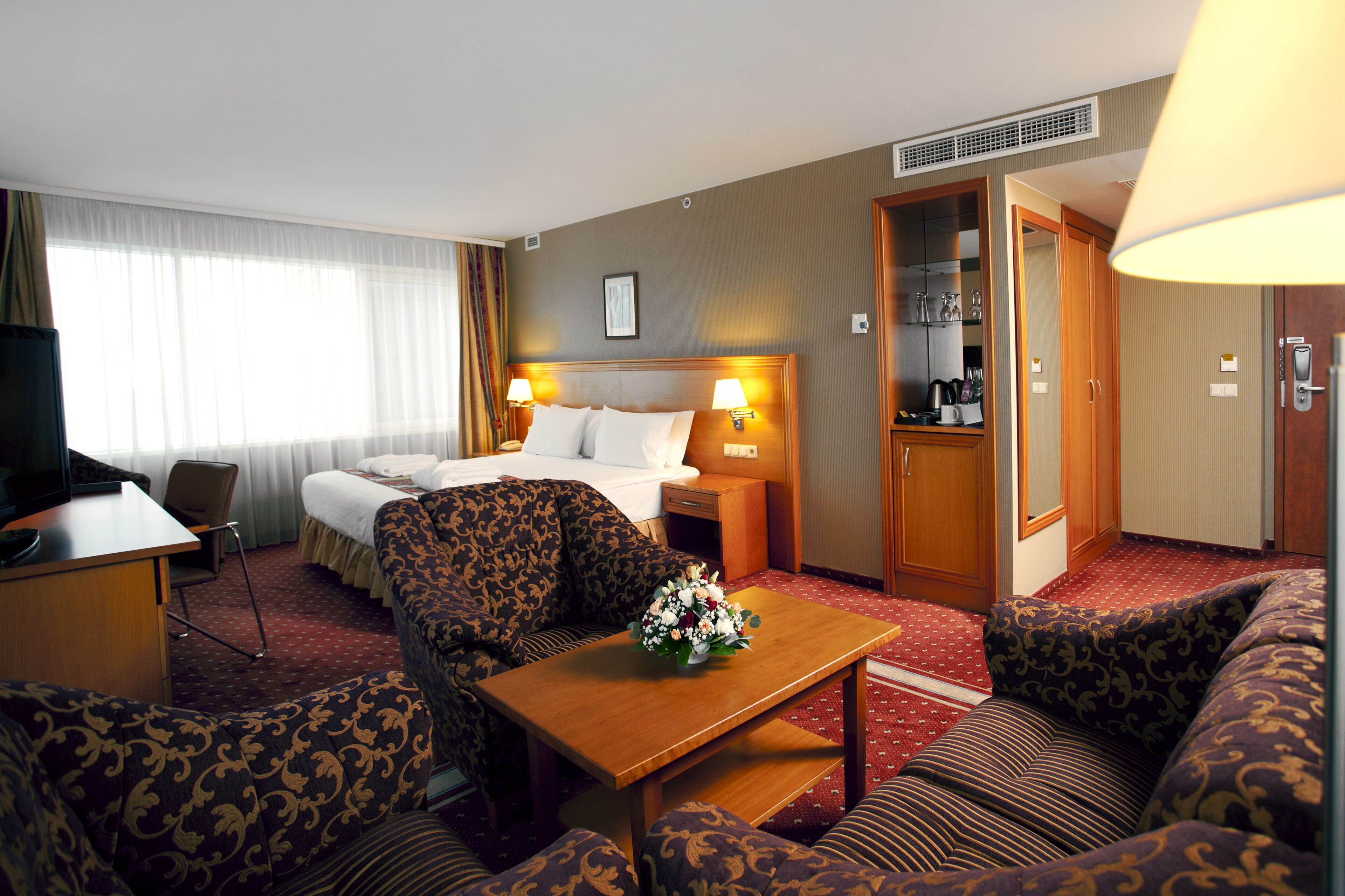 A hotel room with a bed, two sofas, a small table with a flower arrangement, a desk, and a wardrobe.
