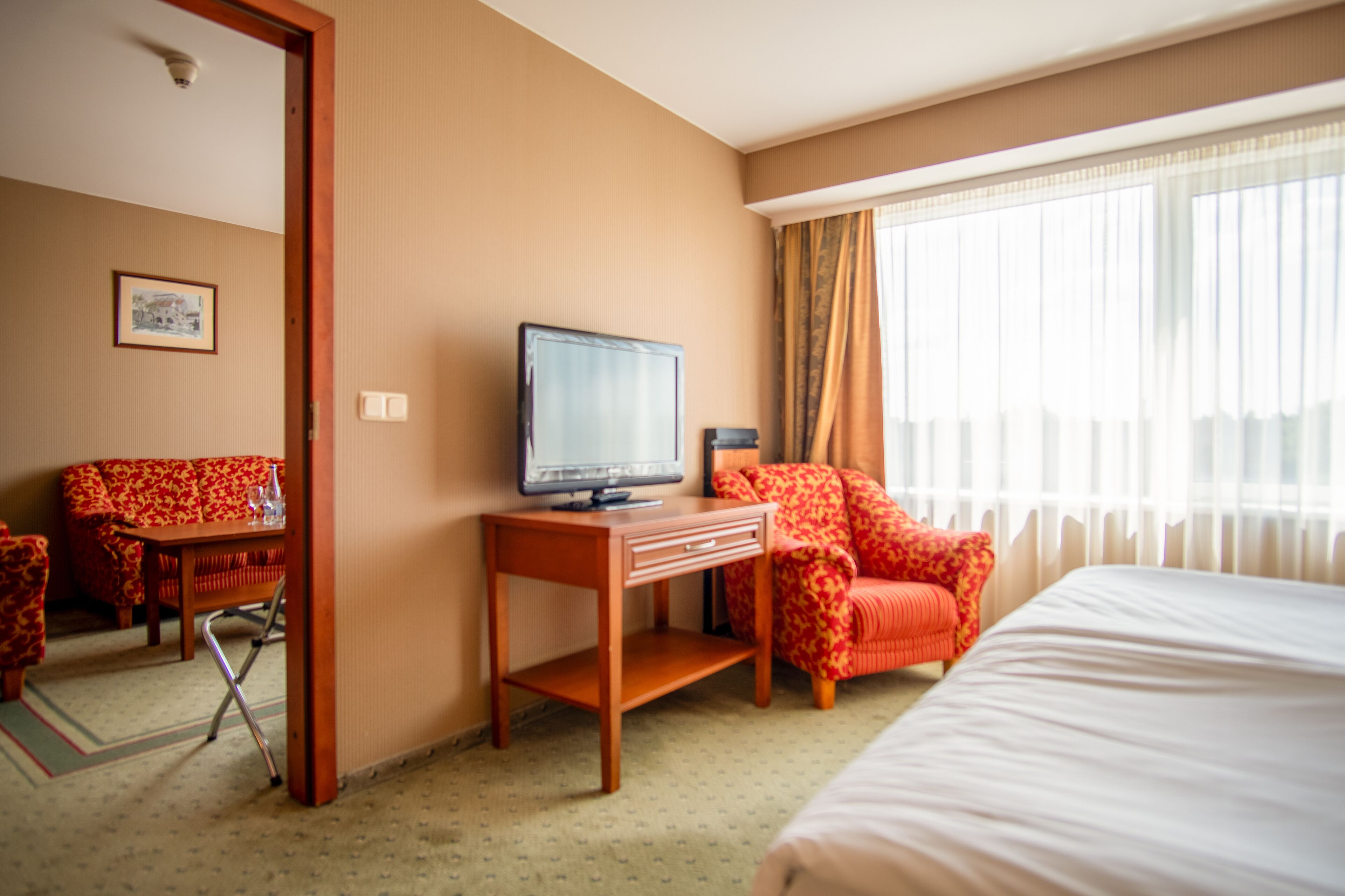 Una habitación de hotel con cama, escritorio con televisor, sillón rojo estampado y zona de comedor con mesa y sillas.