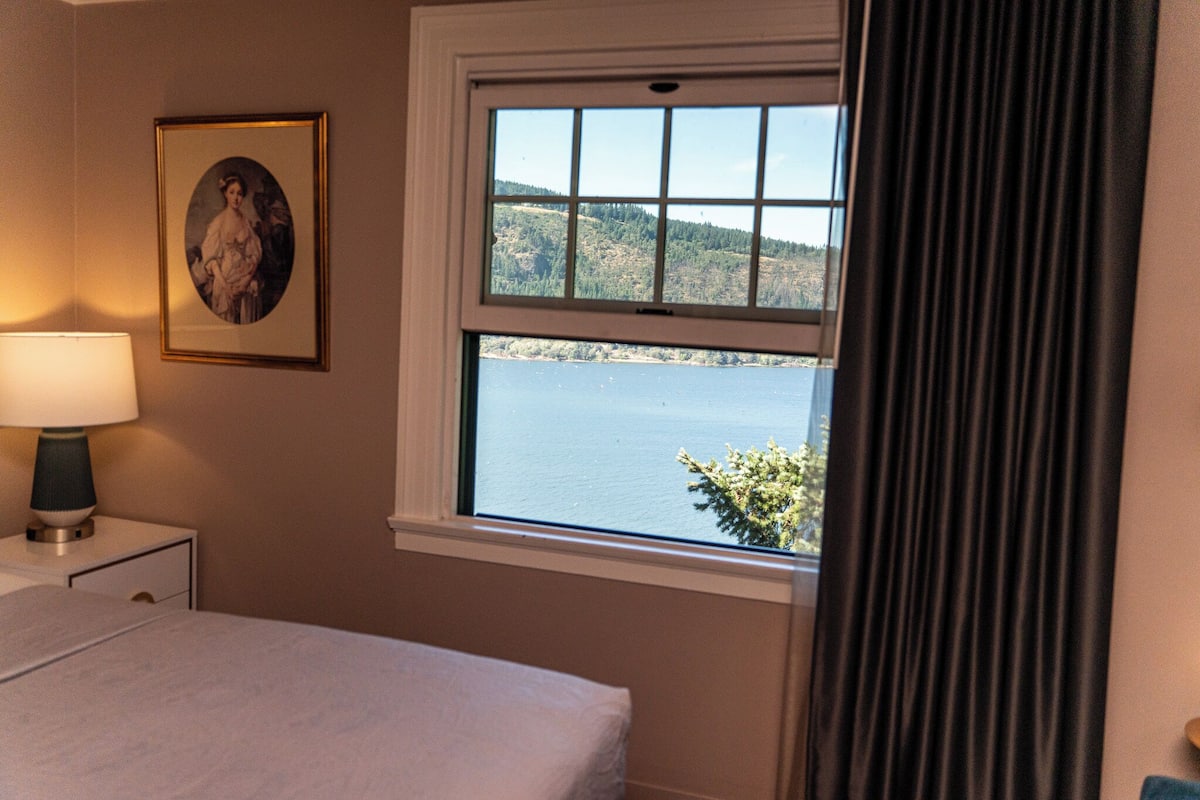 A bedroom with a bed, a nightstand with a lamp, a framed picture on the wall, and a window with a view of a lake and hills.