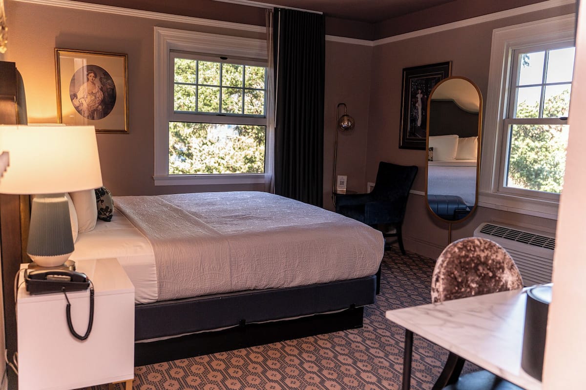 A hotel room with a bed, bedside table, lamp, chair, and a large mirror.