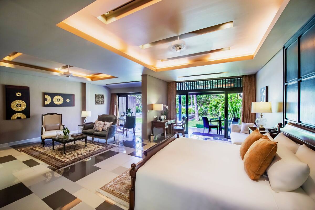 Quarto de hotel espaçoso com uma cama grande, área de estar com sofá e poltronas, escrivaninha e vista para o verde através de amplas janelas.