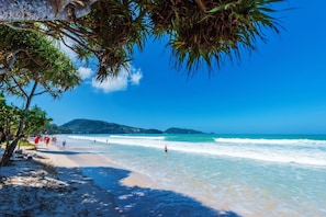 A beach with clear blue water, a sandy shore, and a few people walking.