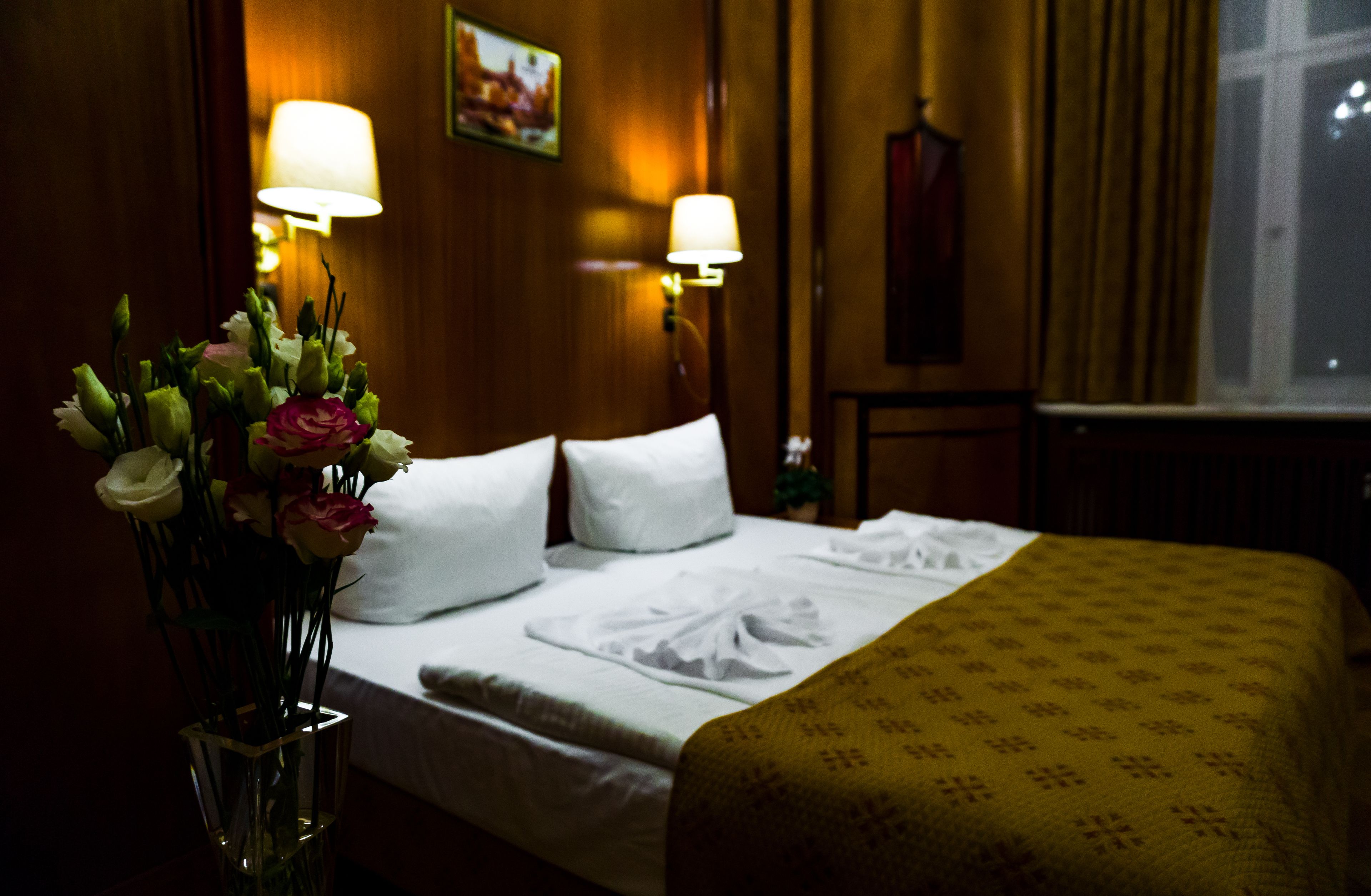 A hotel room with a neatly made bed, a vase of flowers, and two wall-mounted lamps.