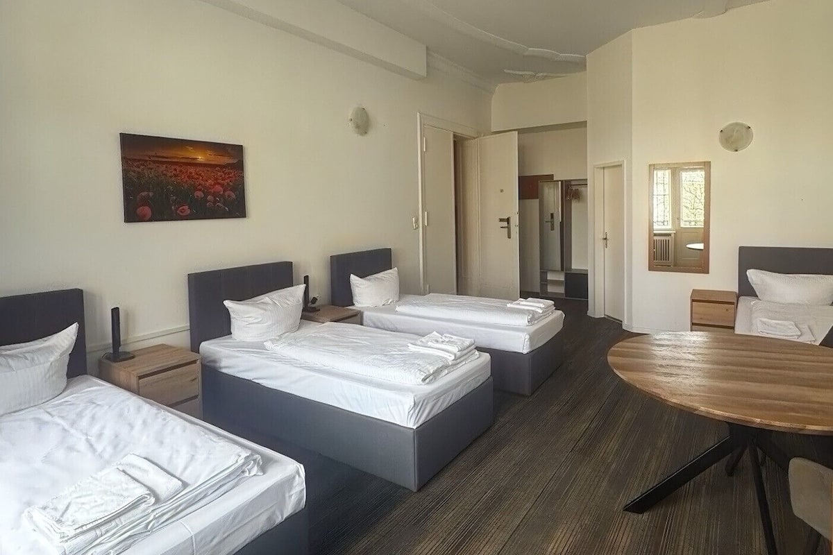 A hotel room with three beds, a wooden dining table, and a painting on the wall.