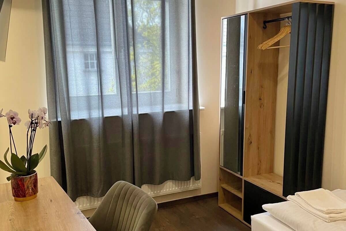 A room with a bed, a wooden table, a chair, a wardrobe, and a window with curtains.