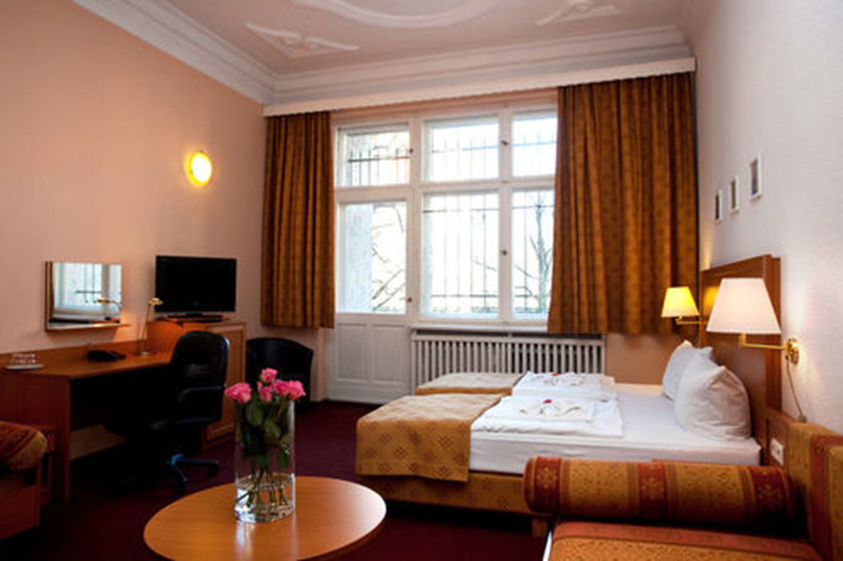 Une chambre d’hôtel comprenant un lit, un bureau avec une chaise, une télévision, une fenêtre avec des rideaux et un vase de fleurs posé sur une table.