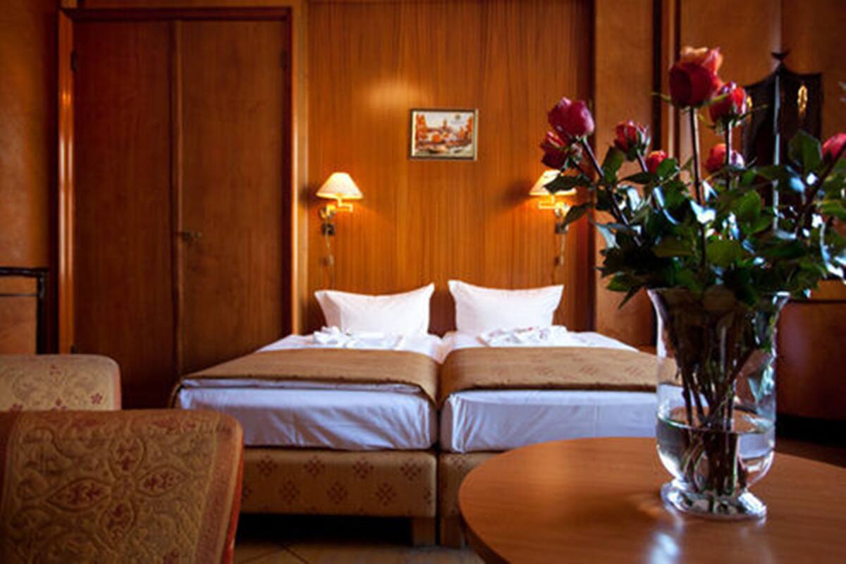 A hotel room with two beds, a wooden door, a wall-mounted lamp, and a vase with flowers on a table.