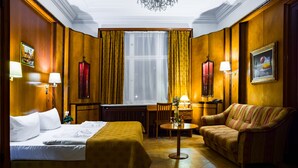 A hotel room with a bed, a sofa, a desk, and a chair. There are two lamps on the walls, a window with curtains, and a chandelier on the ceiling. - Hotel Aster (Berlin)
