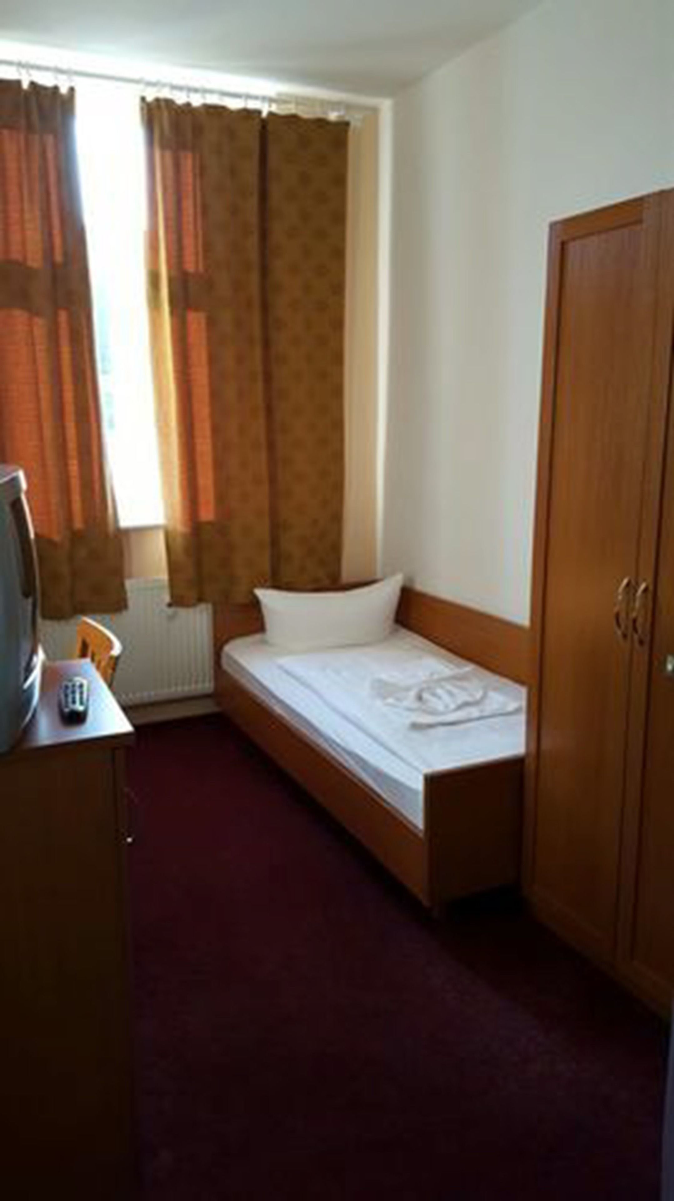 A small, single-bed room with a wooden wardrobe, a desk with a telephone, and a window with curtains.