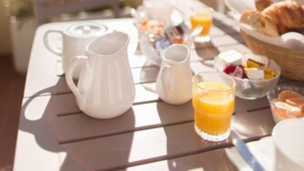 Mesa de café da manhã com jarra, regador e copo de suco de laranja, montada em varanda com corrimão e edifícios ao fundo.