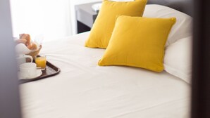 Uma cama bem arrumada com roupa de cama branca e duas almofadas amarelas vibrantes, um tabuleiro com croissants e sumo de laranja, e um candeeiro branco numa mesa de cabeceira.
