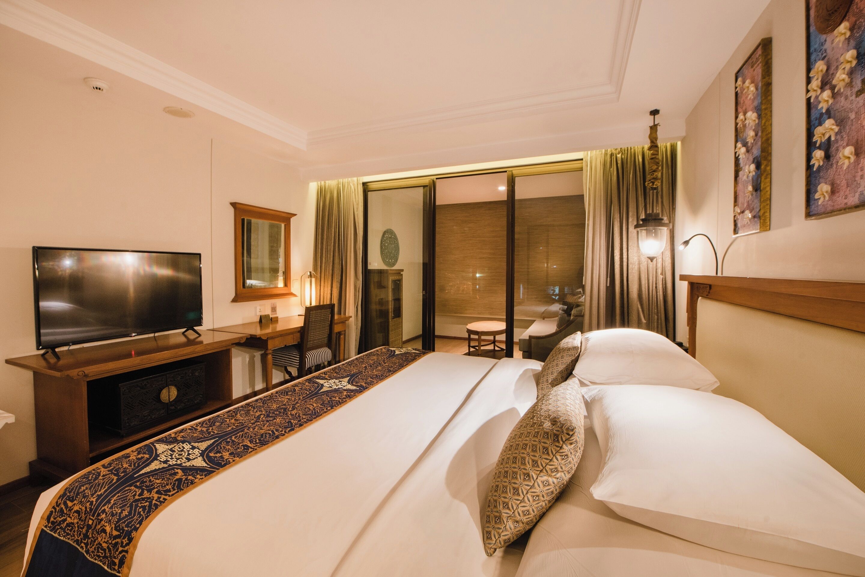A hotel room with a large bed, a flat-screen TV, a desk with a chair, and a sliding glass door leading to a balcony.