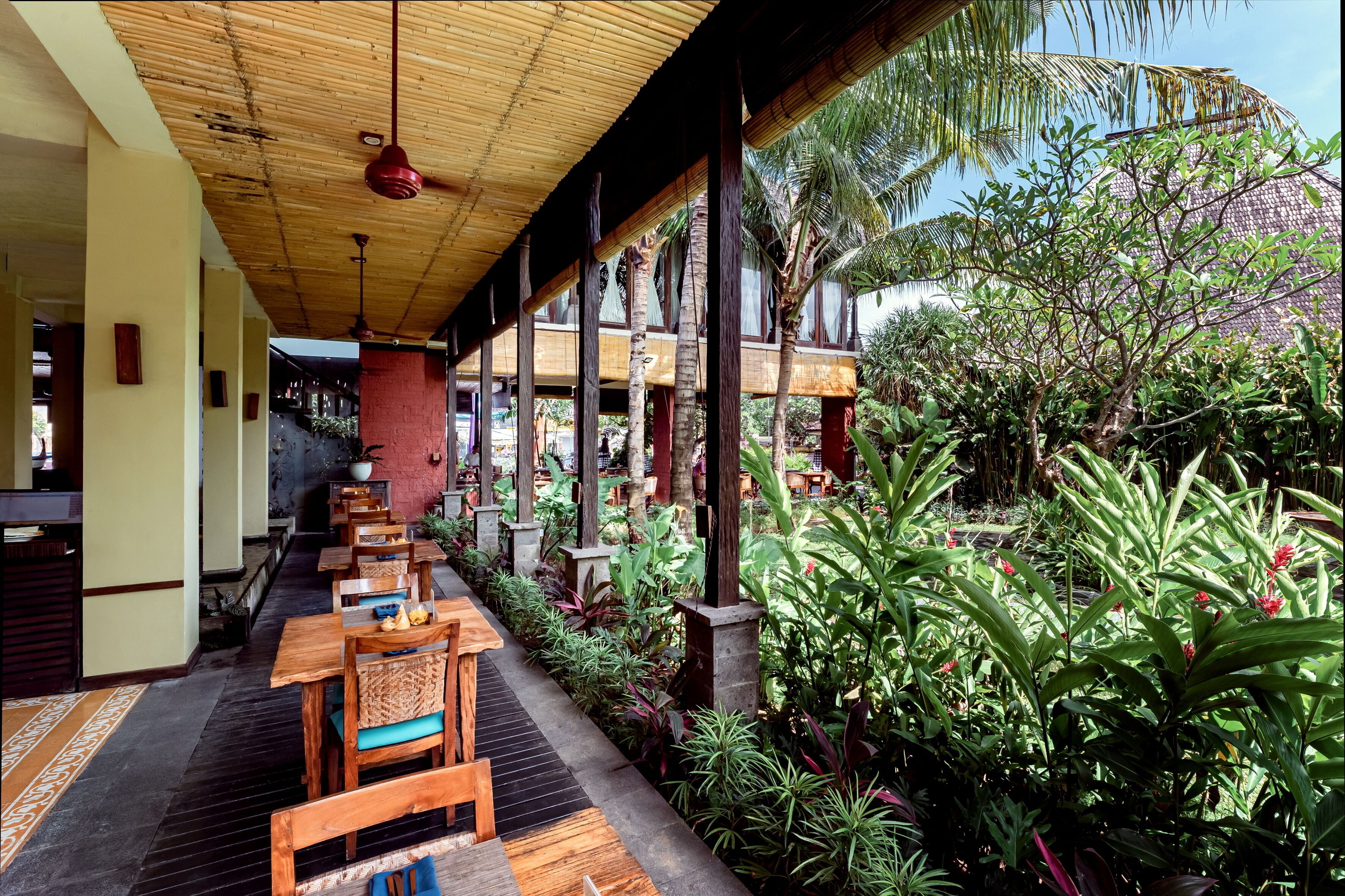 A covered outdoor dining area with wooden tables and chairs, surrounded by lush tropical plants and a bamboo ceiling.