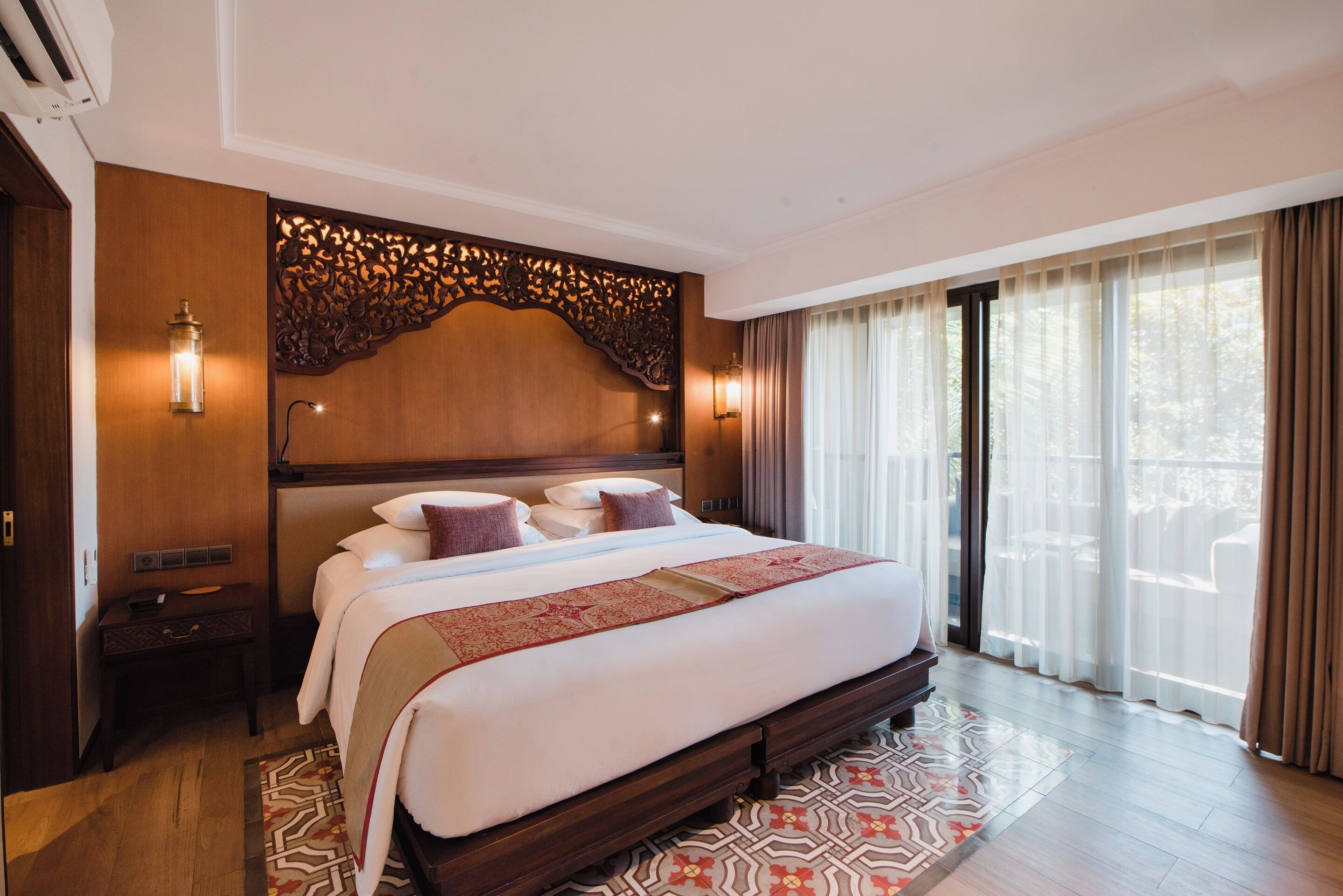 A spacious hotel room with a large bed, wooden headboard, and a view of the outdoors through large windows.