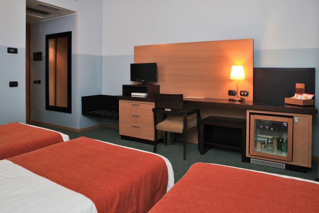 Een hotelkamer met twee bedden, een bureau met een computer, een lamp, een televisie en een minibar.
