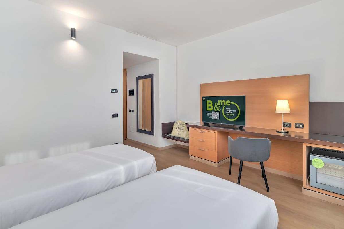 A hotel room with two beds, a desk with a computer, a chair, and a TV displaying a program.