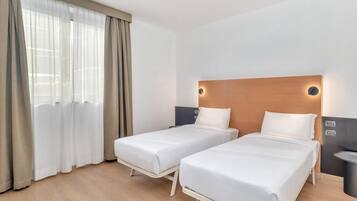 A hotel room with two single beds, a wooden headboard, a bedside table, and a large window with sheer curtains.