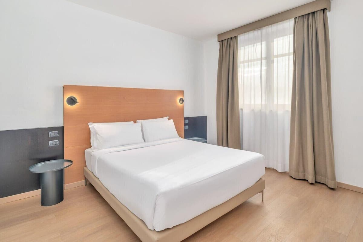 A modern hotel room with a wooden headboard, a large bed, a bedside table, and a window with curtains.