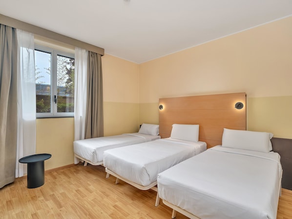 Habitación de hotel con dos camas individuales, una mesita redonda pequeña, una ventana con cortinas y un cabecero de madera con luces fijadas a la pared.