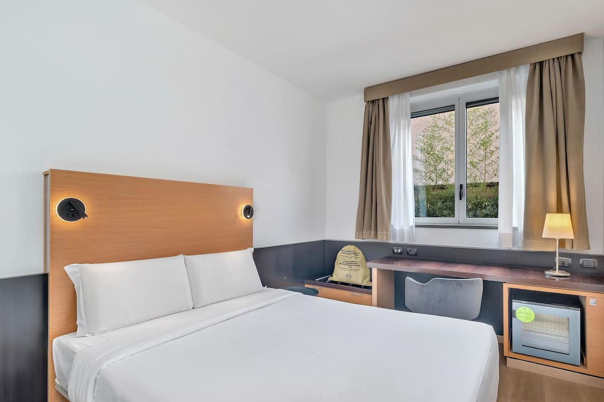 A hotel room with a bed, a desk, a chair, a lamp, and a window with curtains.