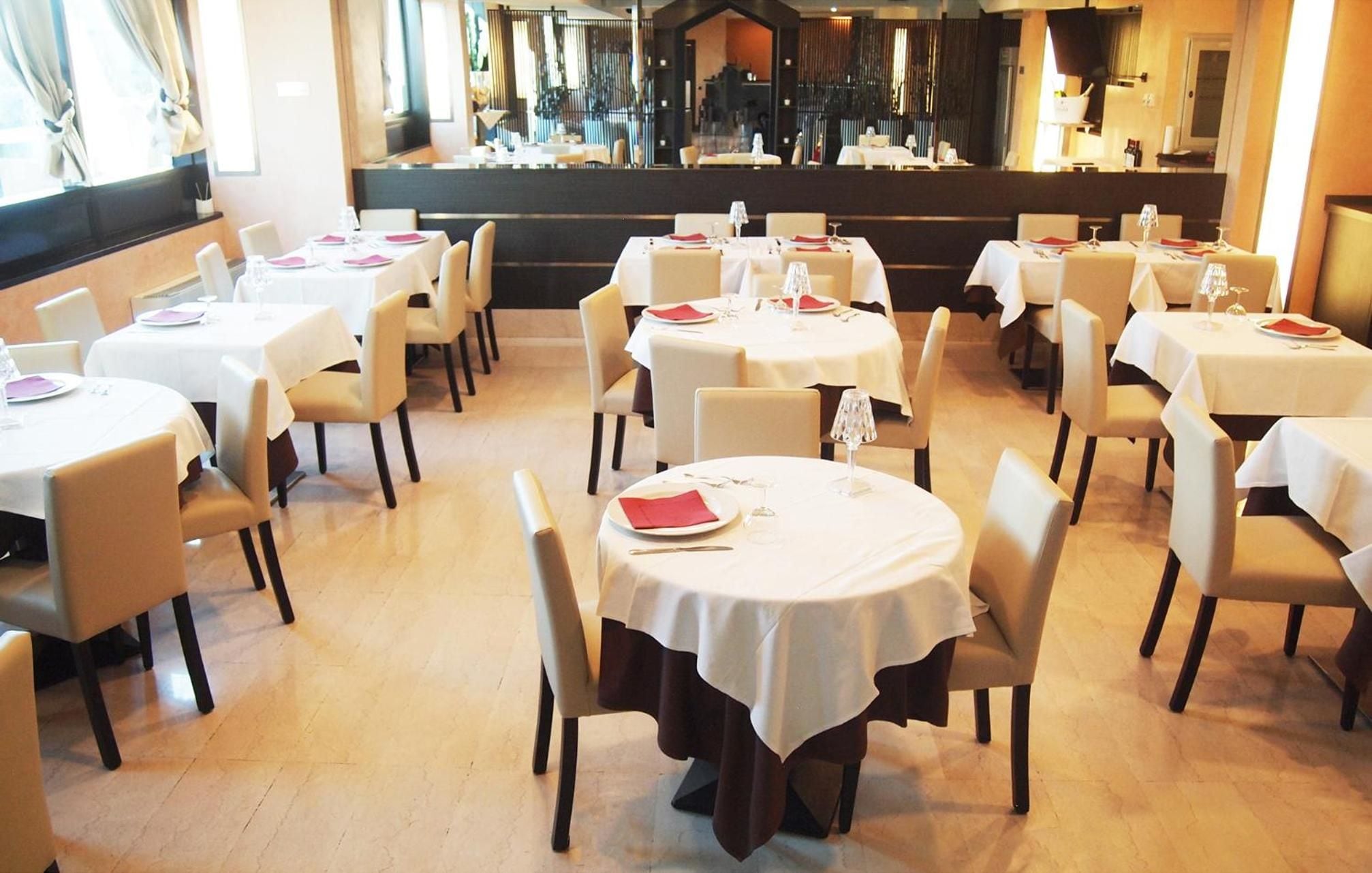 Un restaurante con mesas y sillas redondas, cada conjunto con manteles blancos y servilletas rojas.
