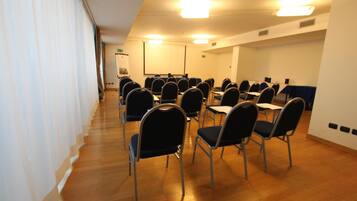 Une salle de conférence avec des rangées de chaises et de tables, un écran de projection et un tableau blanc.