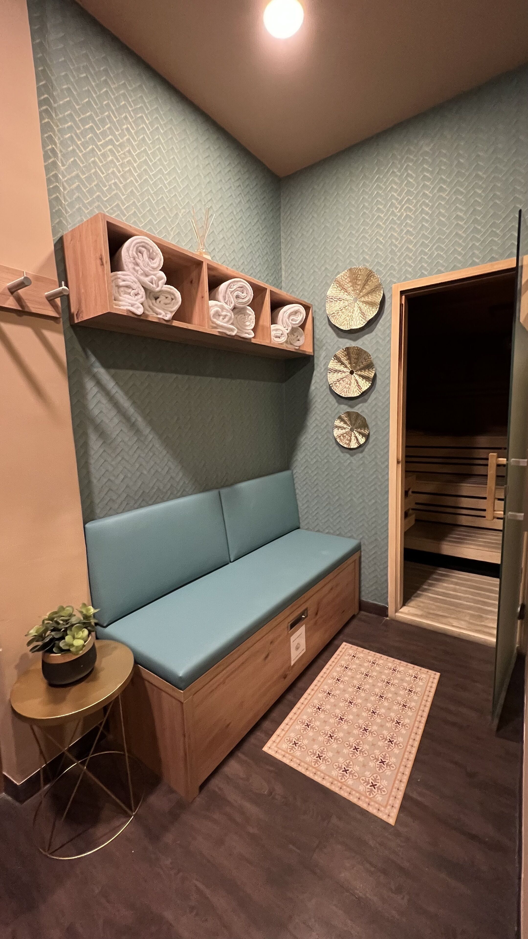 A corner room with a blue cushioned bench, a wooden shelf with rolled towels, a small round table with a plant, and a patterned rug.