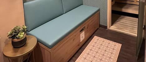A corner room with a blue cushioned bench, a wooden shelf with rolled towels, a small round table with a plant, and a patterned rug.