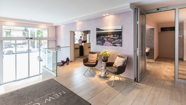 A modern hotel lobby with a reception desk, seating area, and a large window offering a view of the street. - New Orly (Munich)