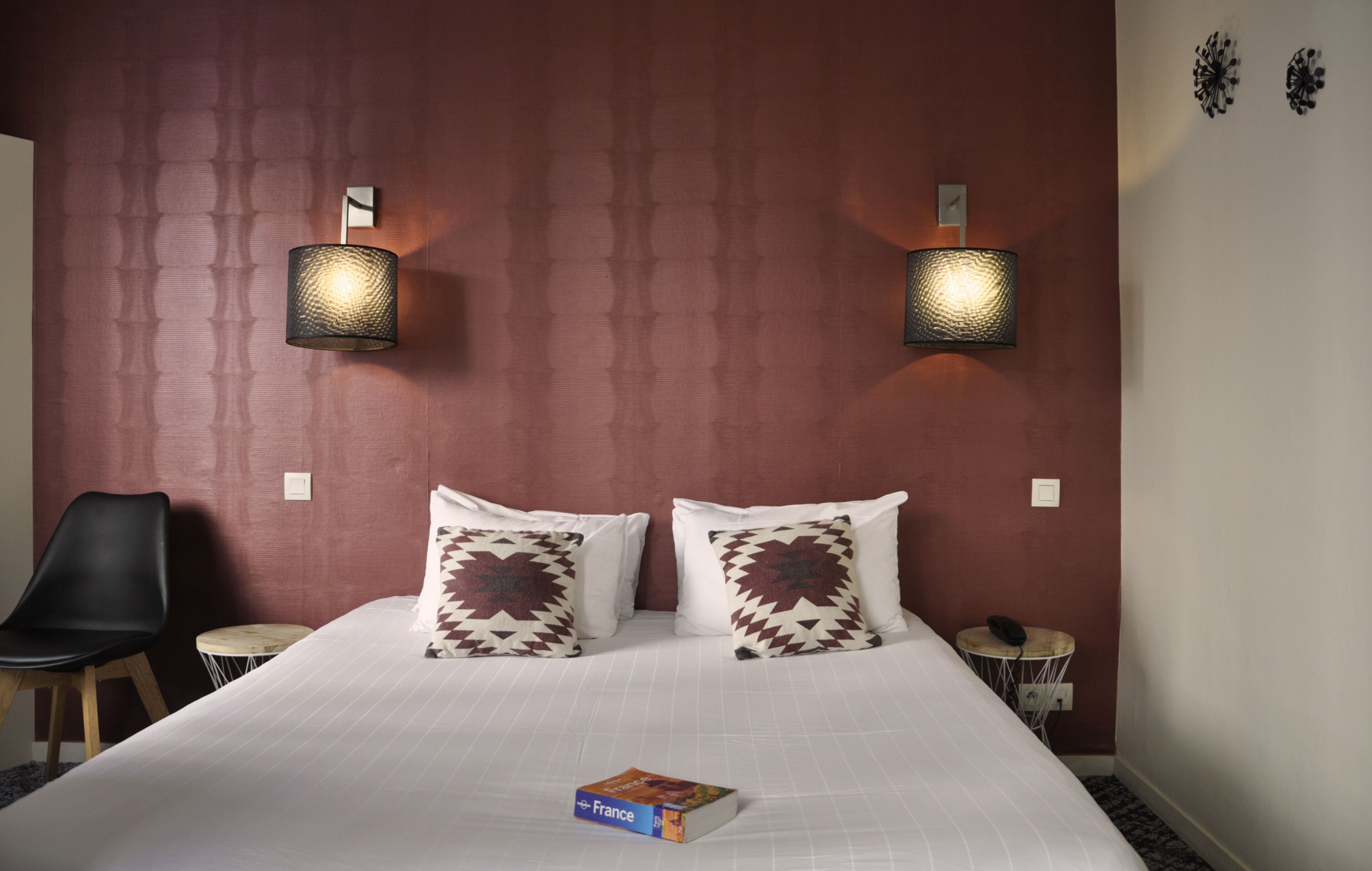 A hotel room with a large bed, two wall-mounted lamps, a black chair, a small round table, and a book titled "France" on the bed.